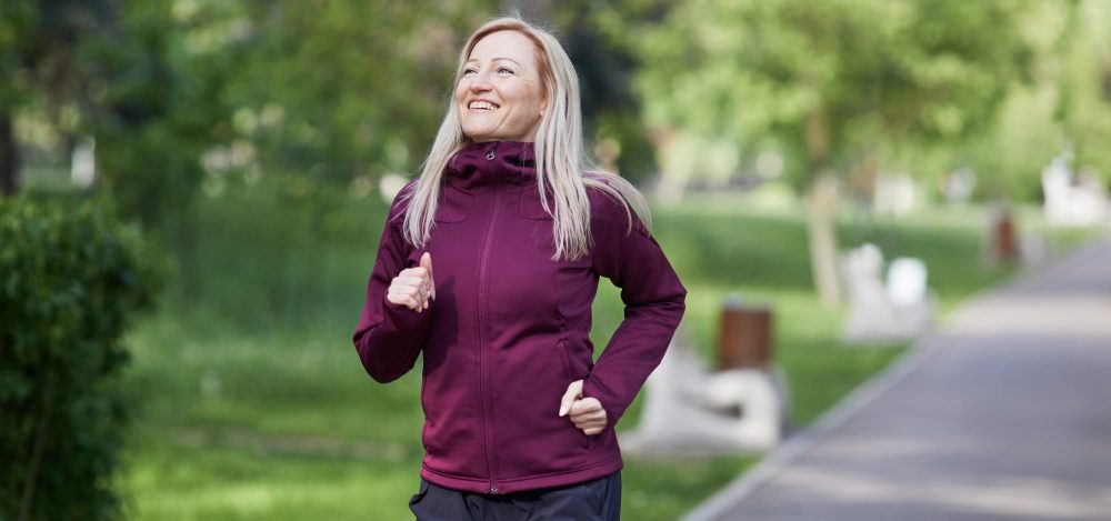 Middle-aged woman enjoys running in a sunlit park, smiling broadly